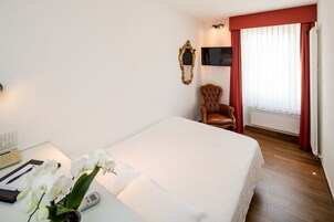 Habitación de hotel con cama, escritorio con teléfono, silla, televisor y ventana con cortinas.