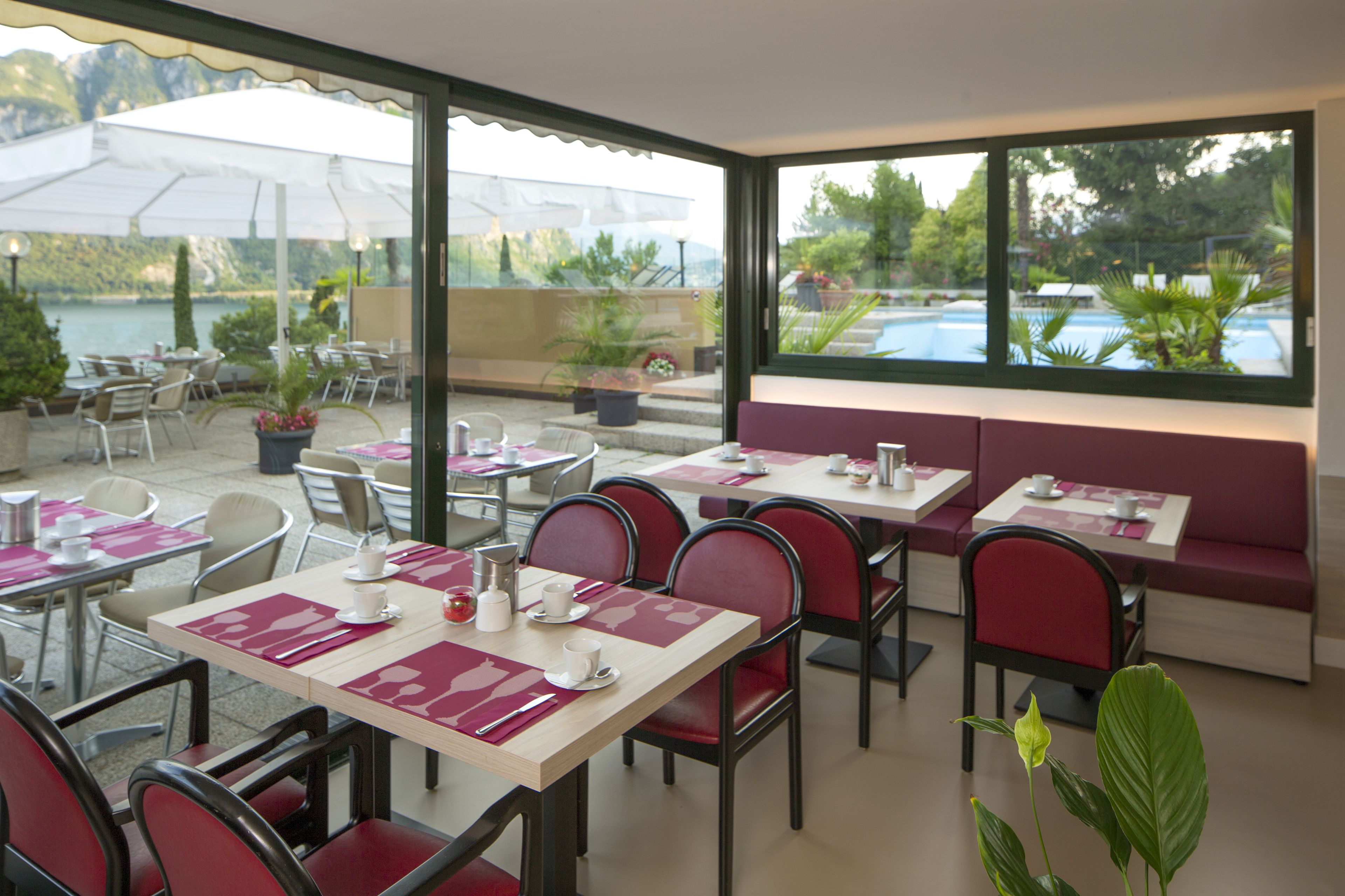 Un restaurante con terraza, mesas con sillas rojas y vistas a un lago y montañas.