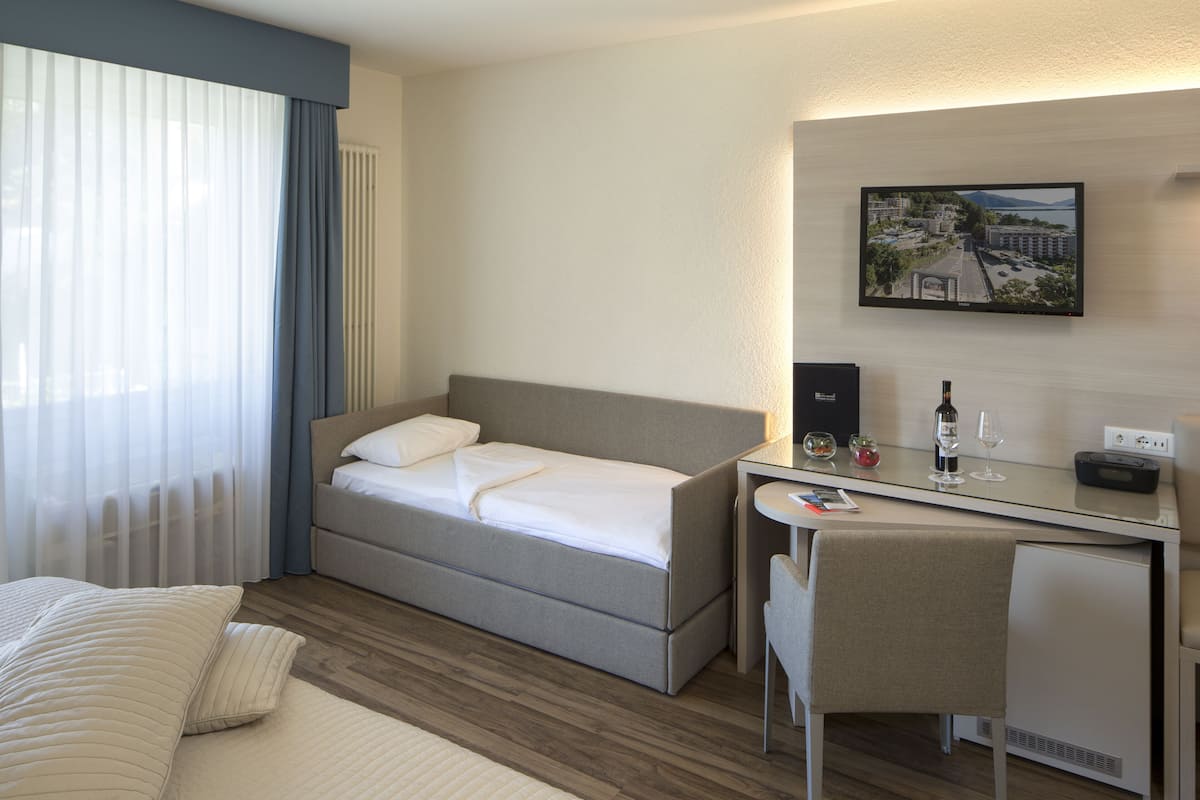 Ein Hotelzimmer mit Bett, Schreibtisch, Stuhl, einem kleinen Tisch, einer Weinflasche, einem Glas, einem Bild an der Wand und einem Fenster mit Vorhängen.