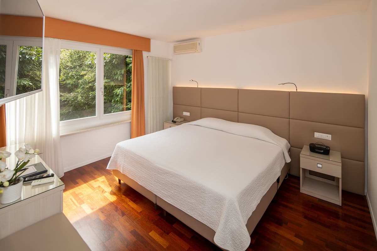 Ein modernes Schlafzimmer mit einem großen Bett, Nachttischen, einem Fenster mit Vorhängen und Blick ins Grüne.