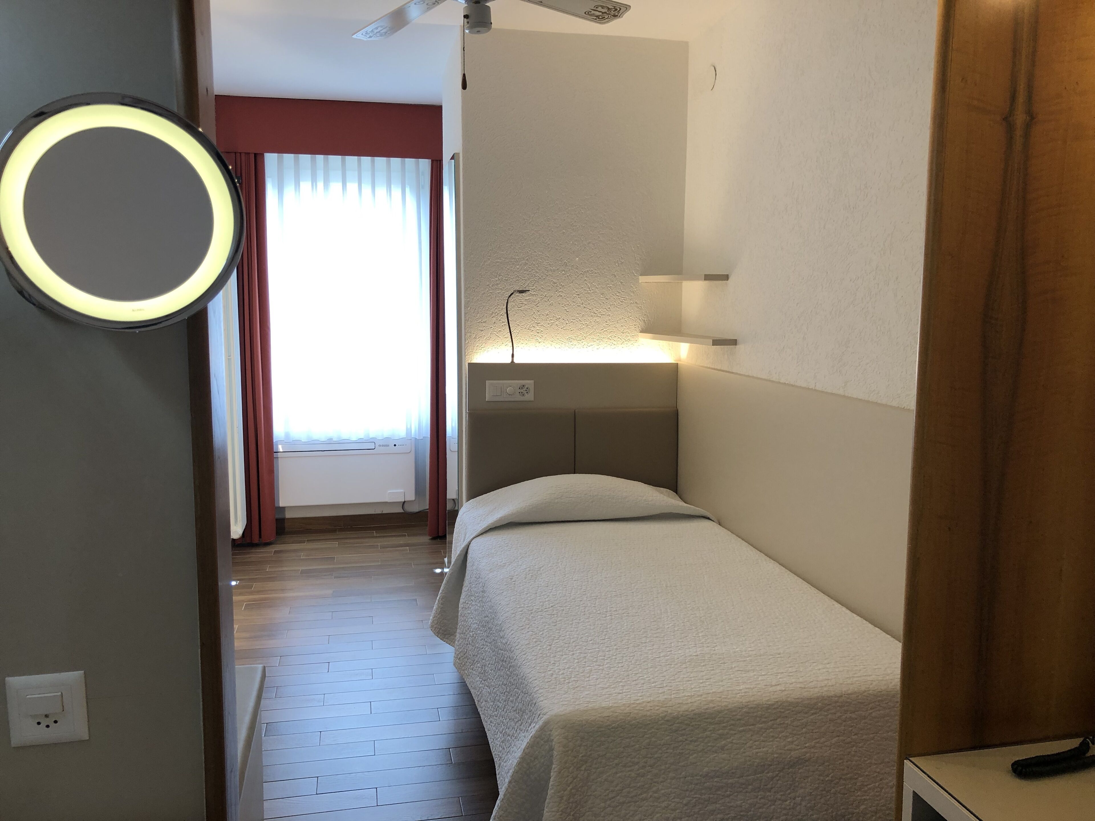 Een hotelkamer met een bed, nachtkastje, spiegel met verlichting, plafondventilator, raam met gordijnen en een radiator.