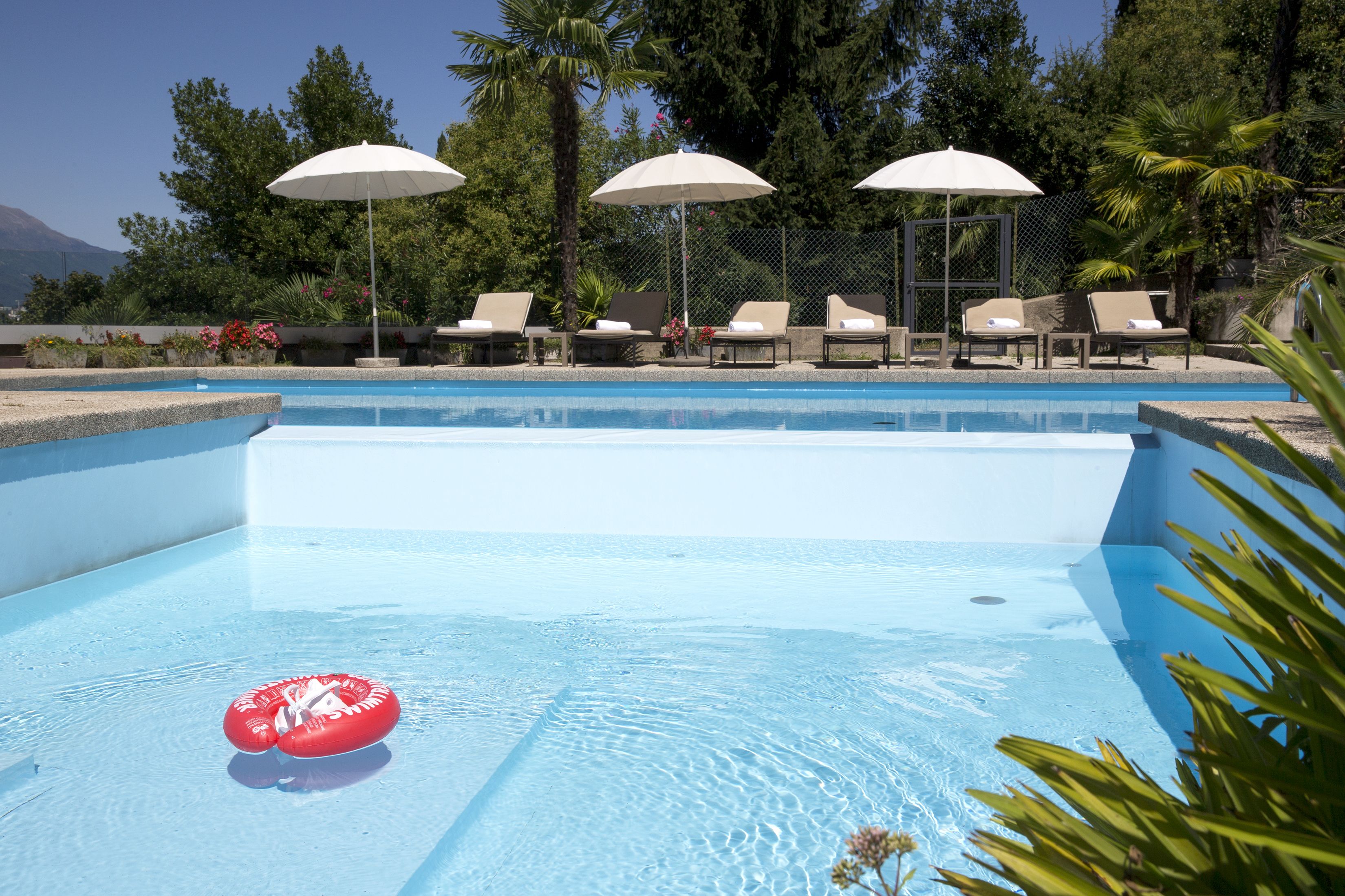 Una piscina con sillas de descanso y sombrillas, rodeada de árboles y una montaña al fondo.