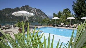 Un área de piscina con sillas de descanso y sombrillas, rodeada de montañas y una masa de agua.