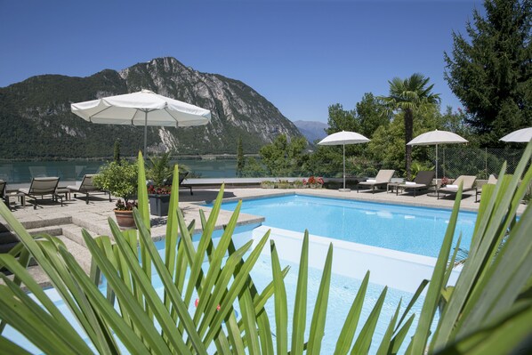 A pool area with lounge chairs and umbrellas, surrounded by mountains and a body of water. - Hotel Campione (Bissone)