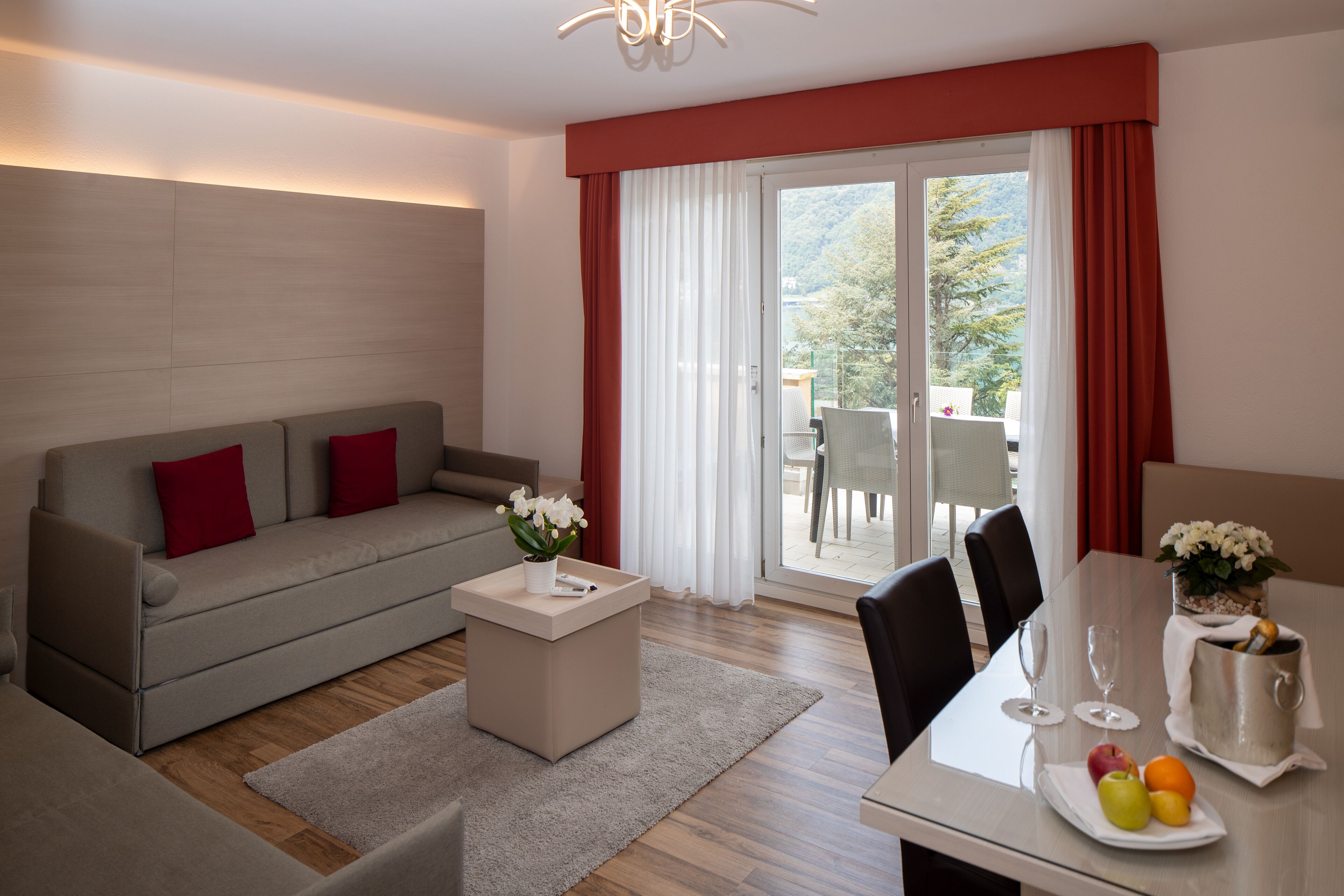 A modern living room with a grey sofa, red cushions, a small table with a vase of flowers, and a dining area with a table set for two.
