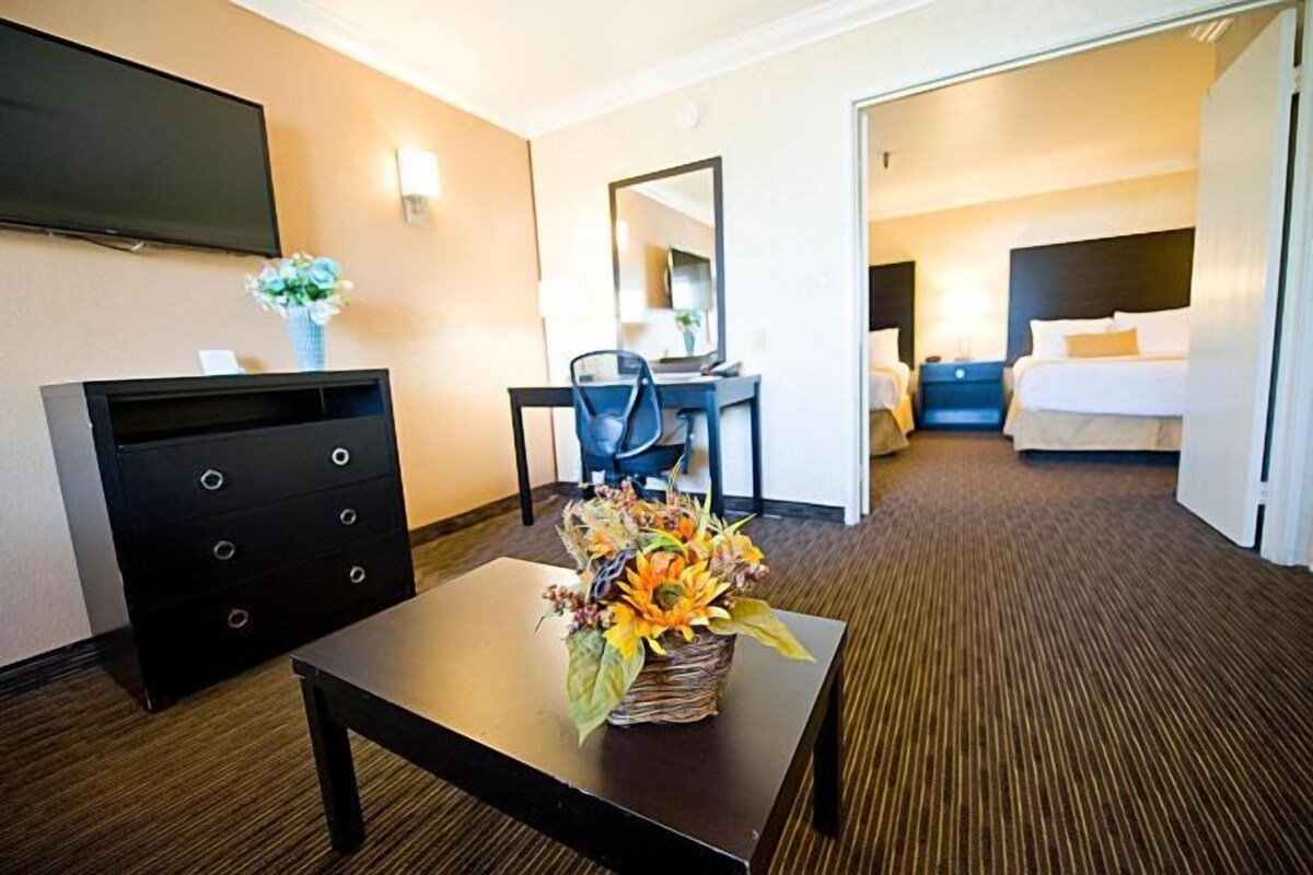 A hotel room with a flat-screen TV, a desk with a chair, a bed with two pillows, and a flower arrangement on a table.