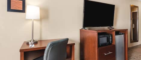 A hotel room with a wooden desk, a black office chair, a flat-screen TV, a microwave, and a refrigerator.