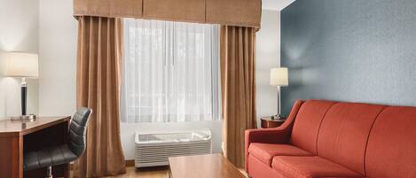 Habitación de hotel con un escritorio de madera, una silla de oficina negra, un sofá rojo, una mesita de madera pequeña y una ventana con cortinas translúcidas.