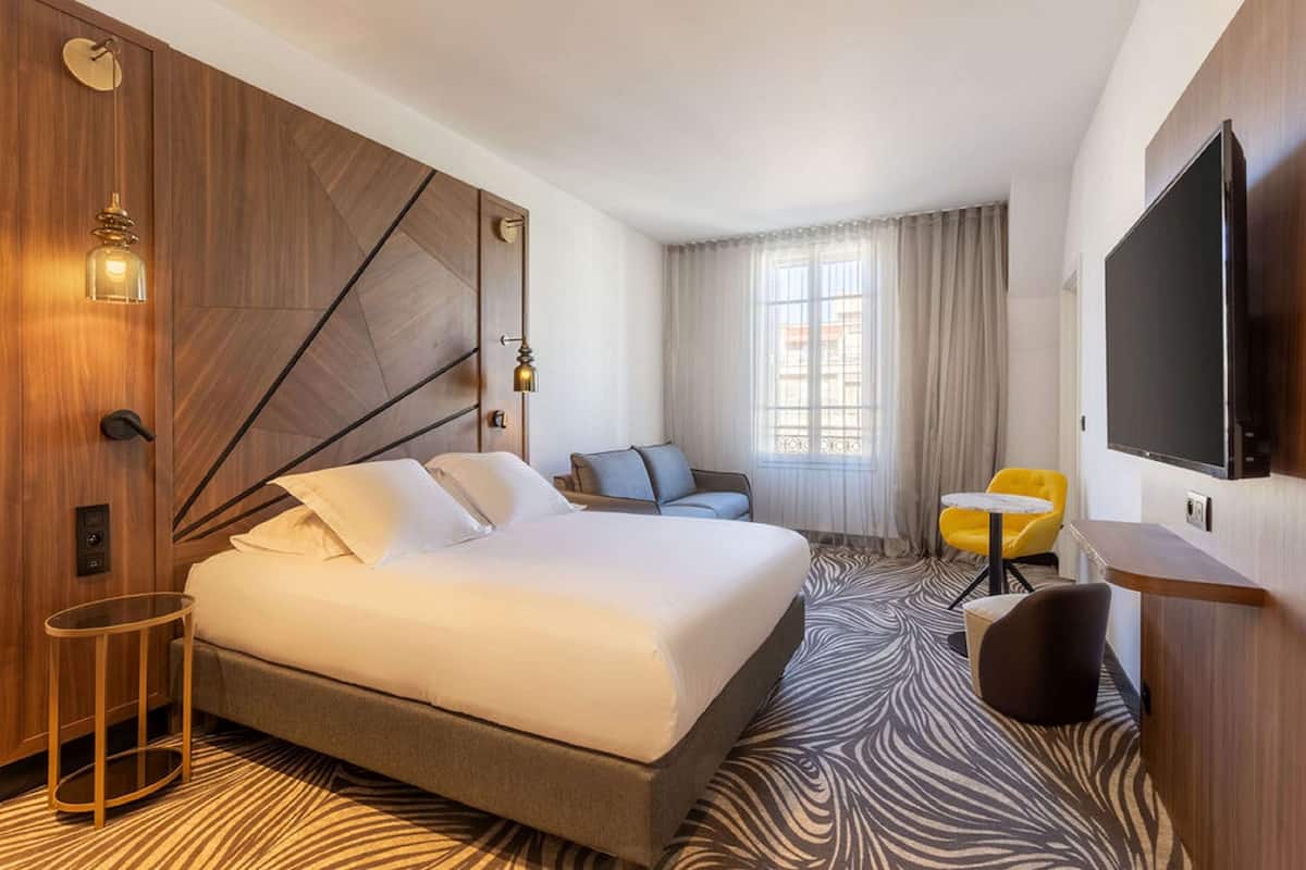 A hotel room with a large bed, a sofa, a small table, a TV, and a window with curtains.