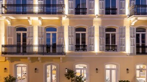 Un edificio de varios pisos con ventanas y contraventanas de marco blanco, balcones con barandillas de hierro forjado y arcos en las entradas.