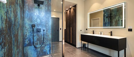 Un baño moderno con un espejo grande, ducha con puerta de cristal y suelo de baldosas oscuras.
