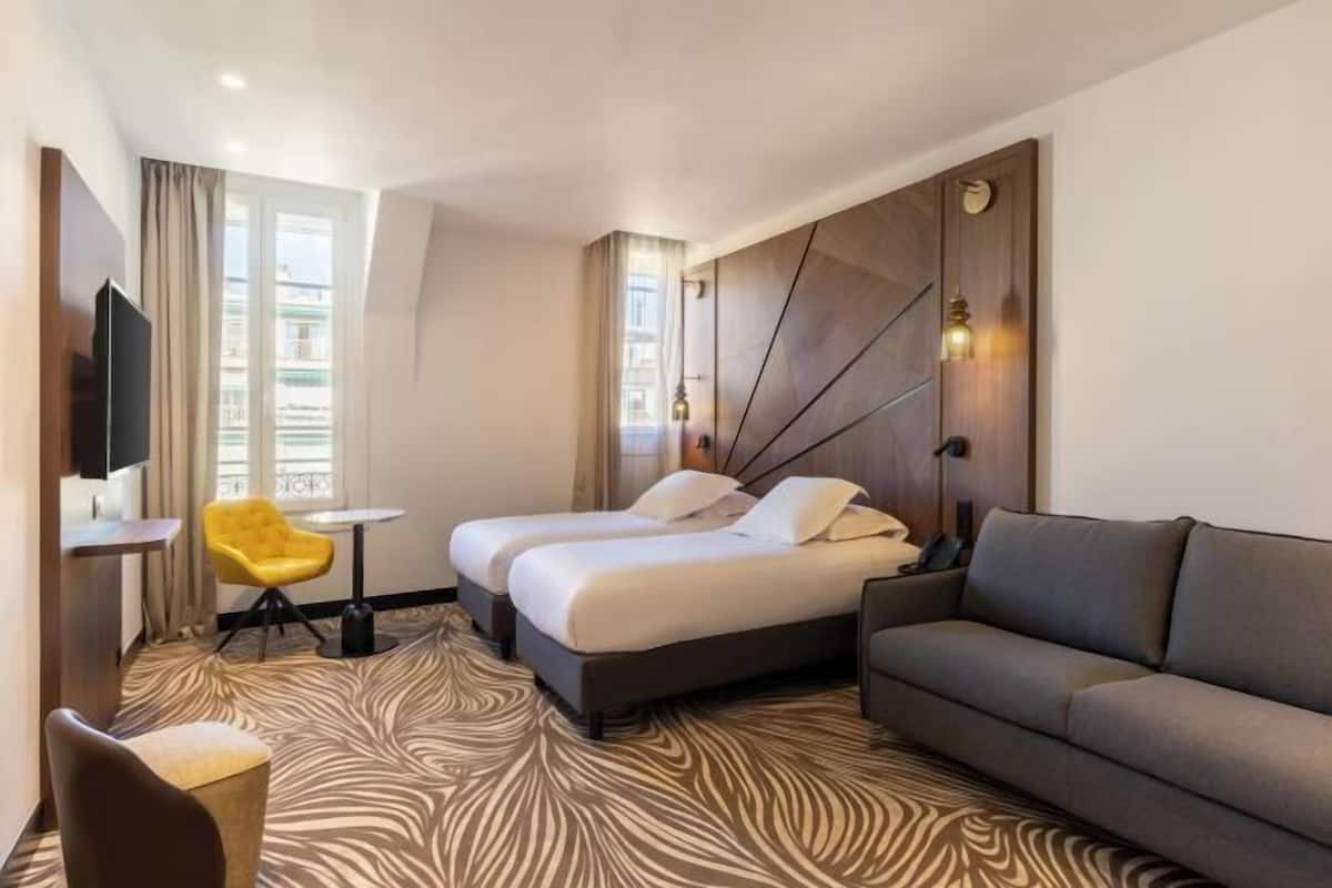 A hotel room with two beds, a sofa, a TV, a small table, a yellow chair, and a patterned carpet.