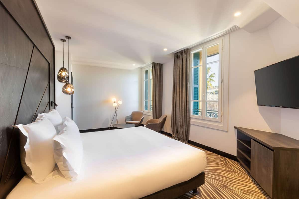 A hotel room with a large bed, a TV mounted on the wall, and a window with curtains.