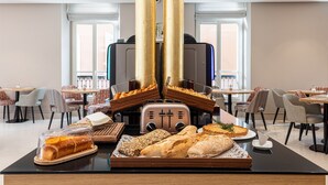 Un bufé de desayuno con panes y pastelería variada, un tostador y zonas de estar con mesas y sillas.
