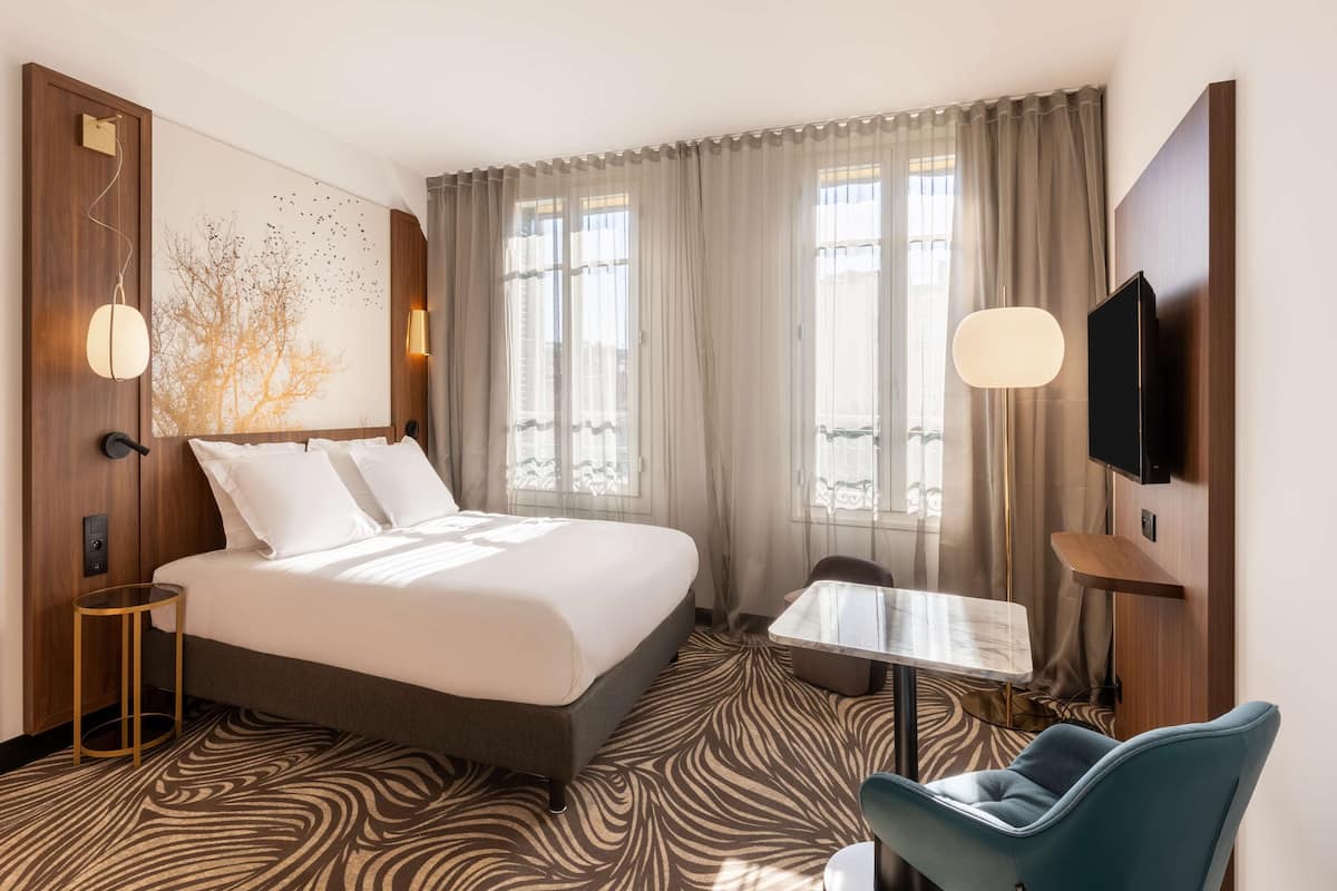 A hotel room with a large bed, a small table, a chair, a TV, and a wall-mounted TV.