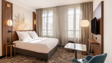 A hotel room with a large bed, a small table, a chair, a TV, and a wall-mounted TV.