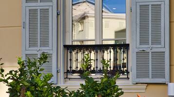 Een raam met witte luiken, een balkon met zwart ijzeren hekwerk en een boom met groene bladeren ervoor.