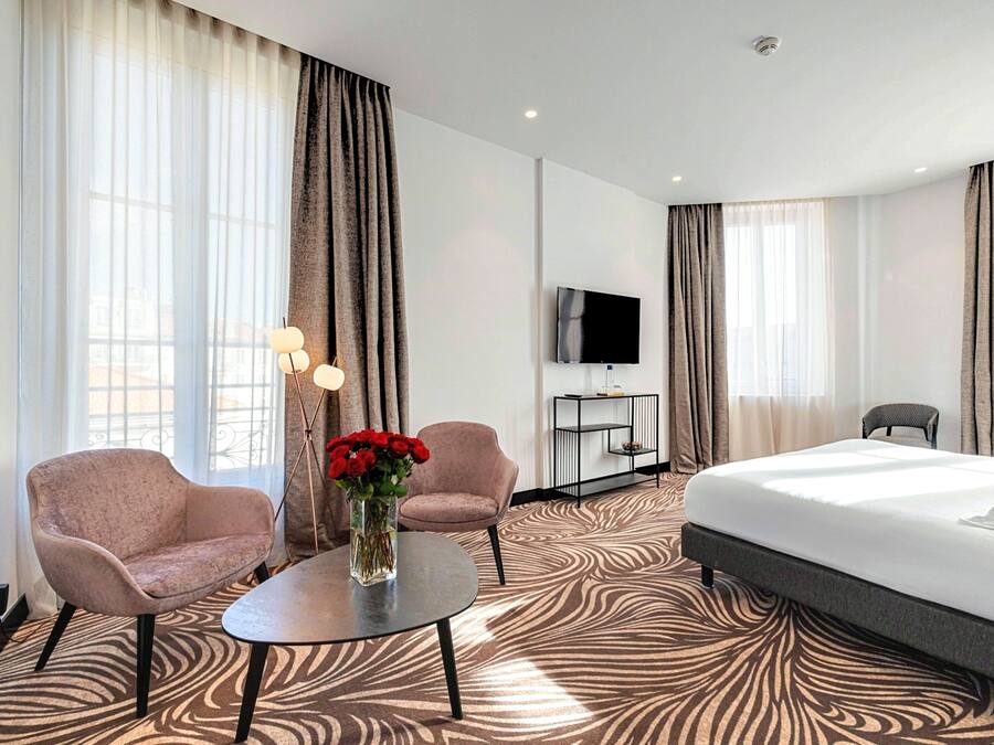 A hotel room with a bed, two armchairs, a small table with a vase of flowers, a TV on the wall, and a patterned carpet.