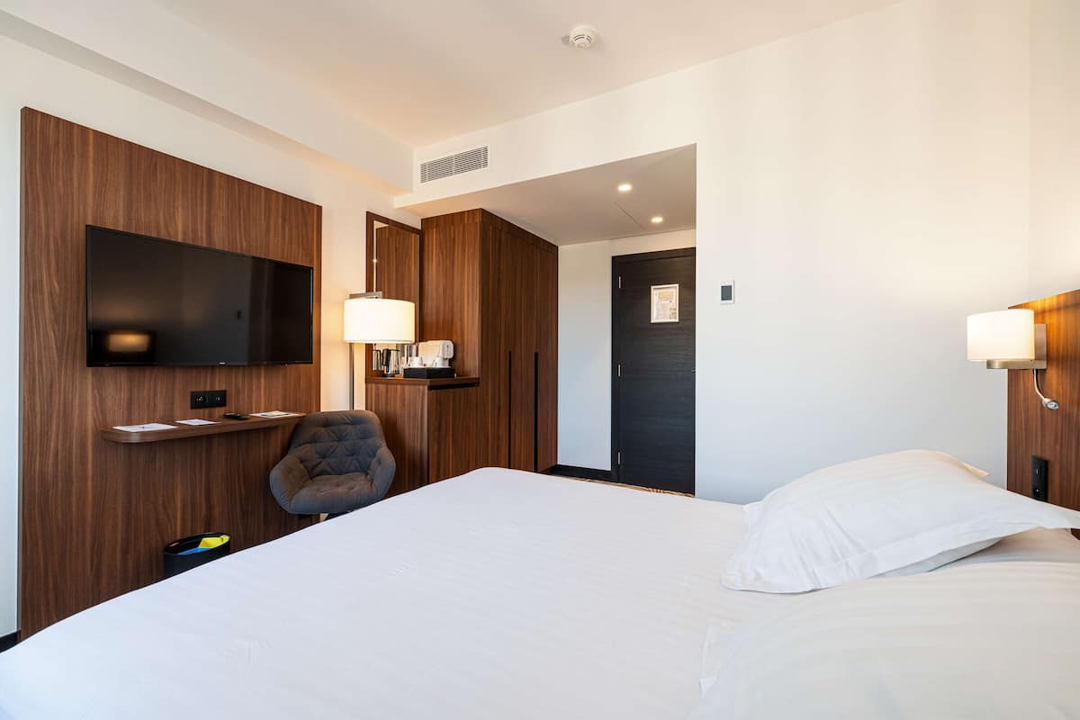 A modern hotel room with a large bed, a flat-screen TV mounted on the wall, a desk with a lamp, and a chair.