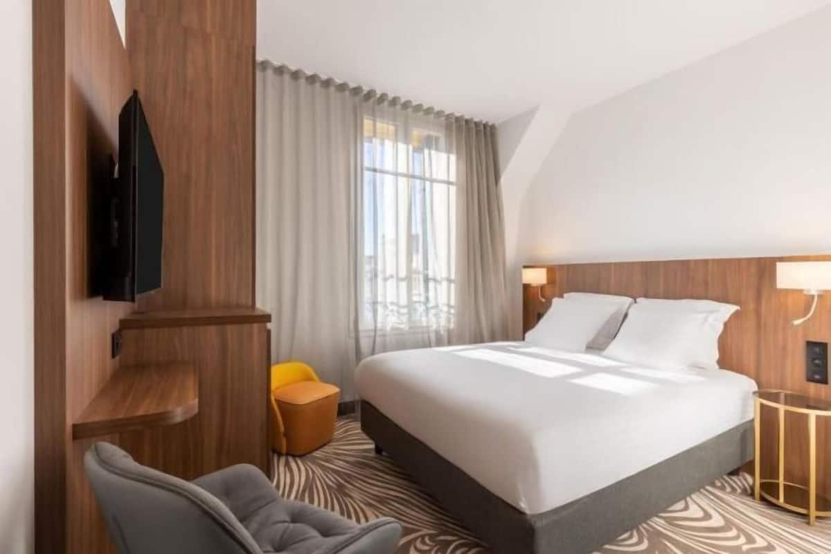 A hotel room with a bed, a TV mounted on the wall, a desk, a chair, and a window with curtains.