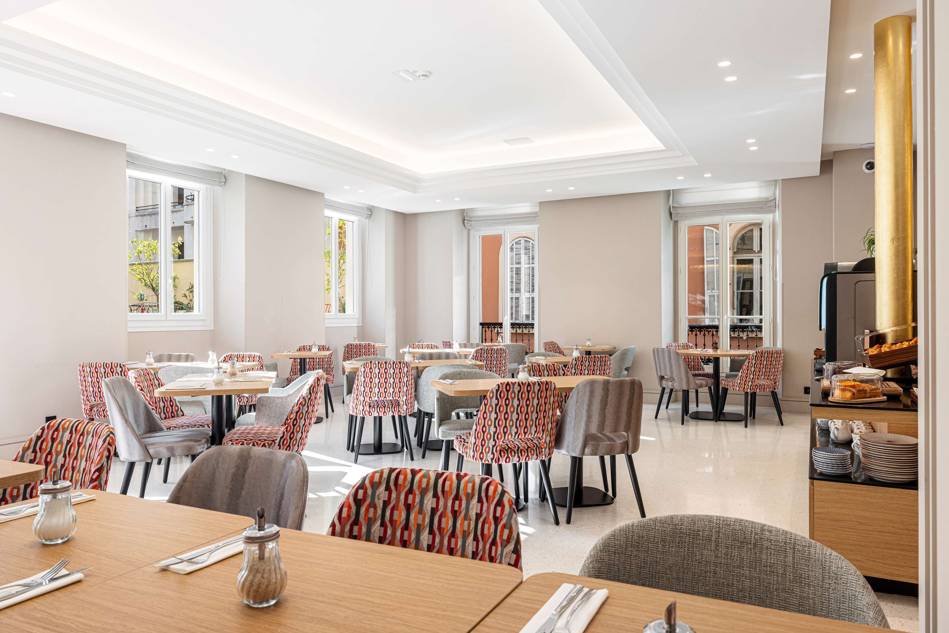 A spacious dining area with multiple tables and chairs, large windows, and a kitchen area visible in the background.
