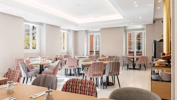 A spacious dining area with multiple tables and chairs, large windows, and a kitchen area visible in the background.