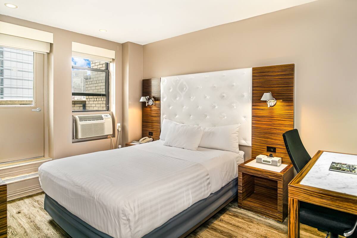 Quarto de hotel com uma cama grande, uma escrivaninha com cadeira, uma luminária na parede e uma janela com persianas.