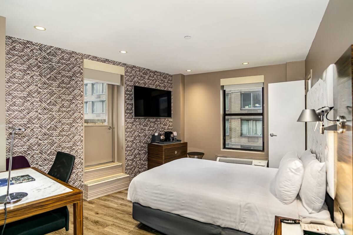 Quarto de hotel com cama, escrivaninha com cadeira, televisão na parede e janela com vista para o exterior.