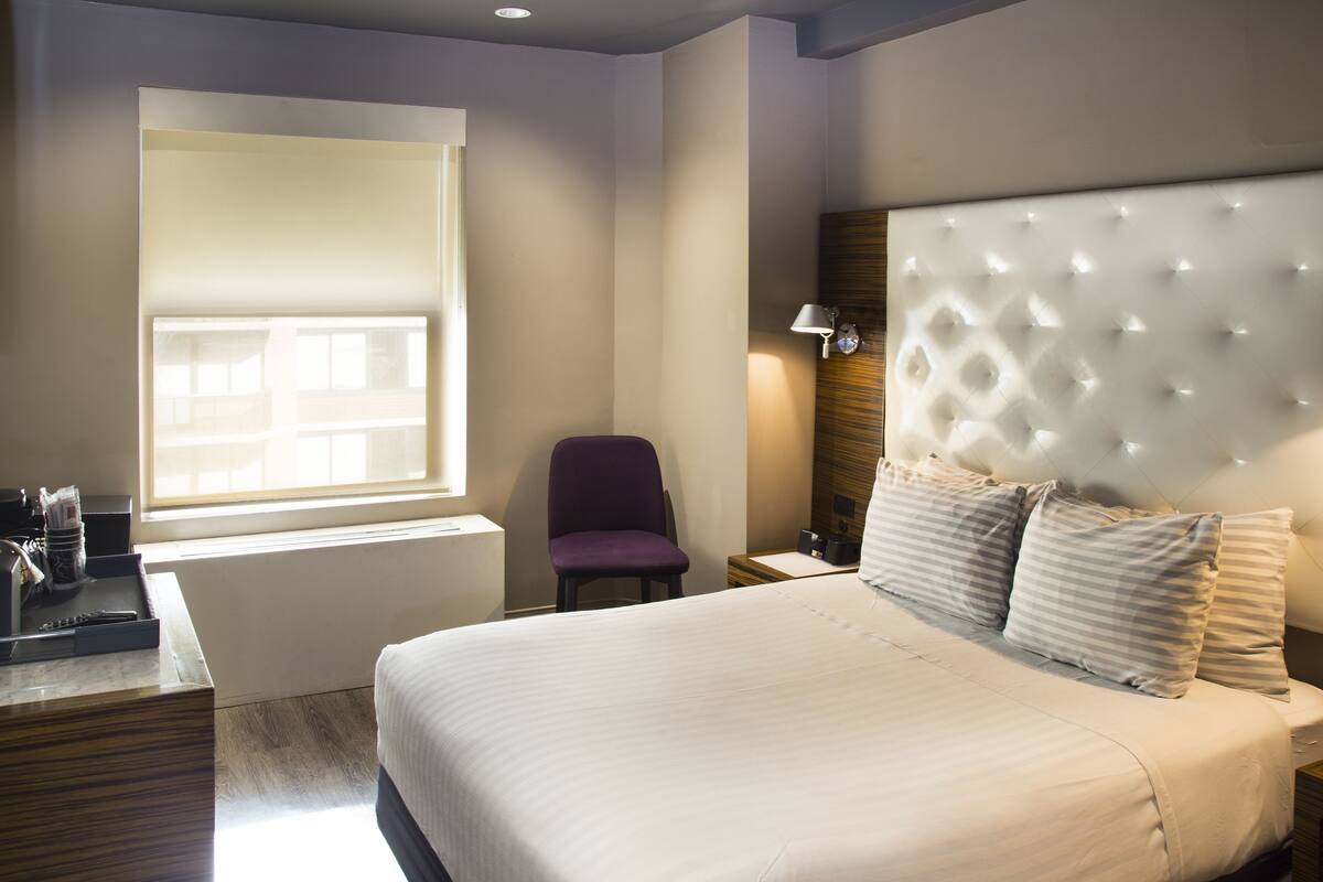 Quarto de hotel com uma cama grande, uma cadeira, uma escrivaninha e uma janela.