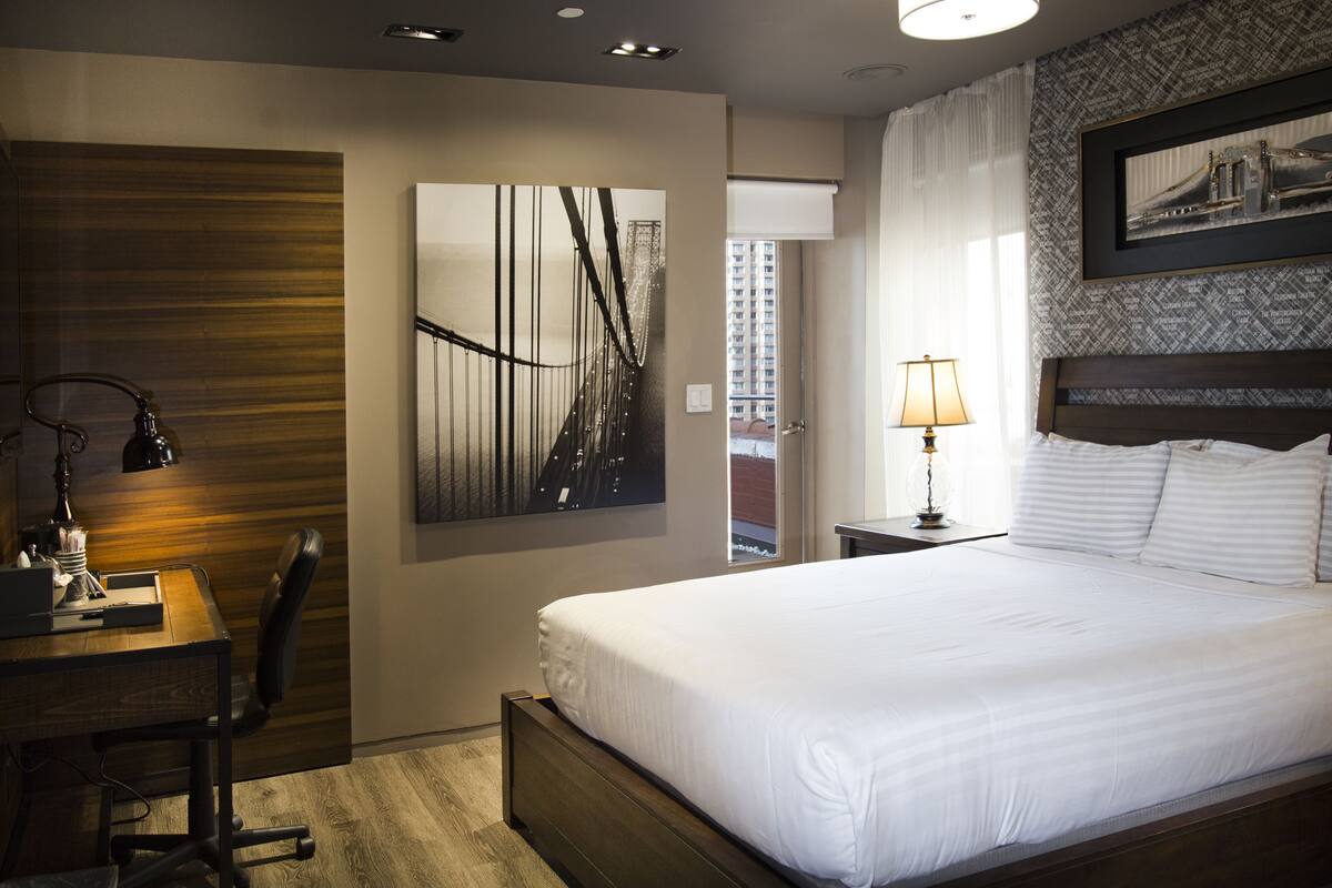 Quarto de hotel com uma cama grande, uma escrivaninha com cadeira, uma luminária e um quadro na parede.