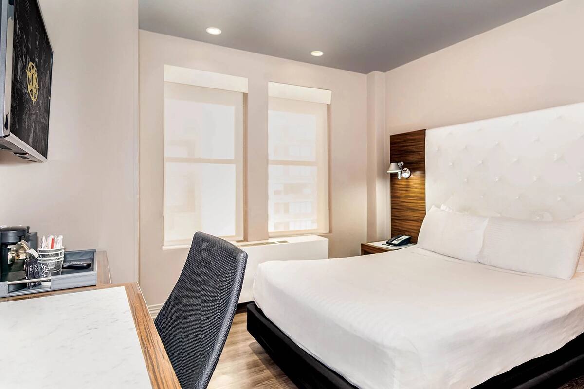 Quarto de hotel com uma cama grande, uma mesa de trabalho com cadeira, mesa de cabeceira e luminária na parede.