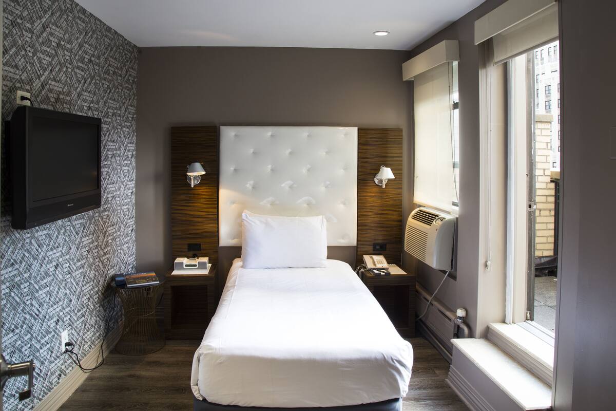 Quarto de hotel moderno com uma cama grande, televisão de tela plana na parede, mesa de cabeceira com abajur e janela com vista para a cidade.
