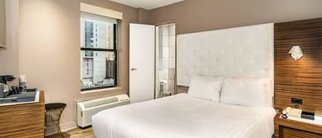 A modern hotel room with a large bed, a desk with a chair, a window with a view of buildings, and a wall-mounted lamp.
