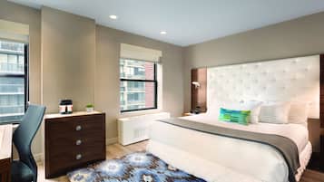 A bedroom with a large bed, a desk, and a chair. There is a window with a view of buildings outside.