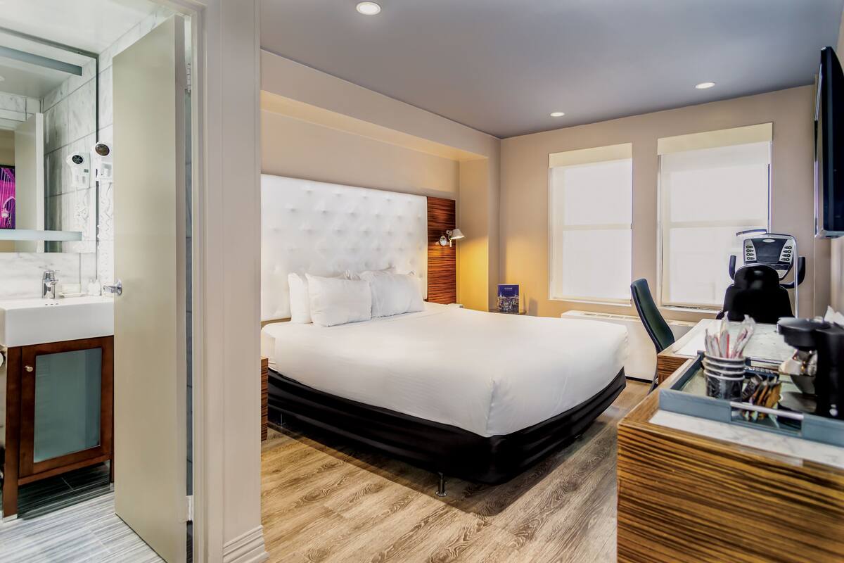 Quarto de hotel moderno com uma cama grande, uma escrivaninha com cafeteira e um banheiro com espelho e pia.