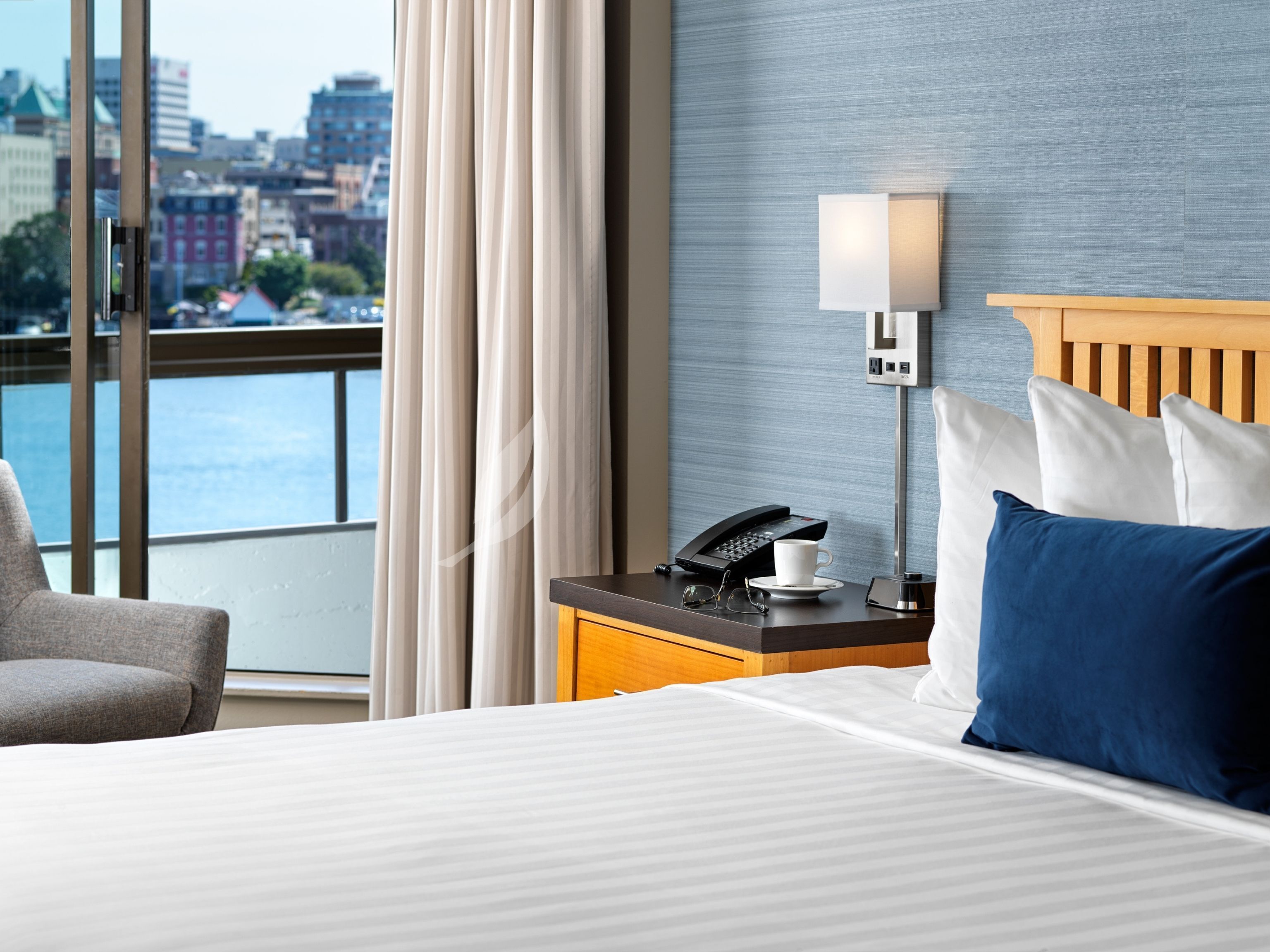 A hotel room with a bed, a nightstand with a lamp, a phone, and a cup. There is a view of a cityscape through a window.