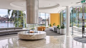 A modern lobby with a central circular seating area, a fire feature, and large windows offering a view of the city and river.