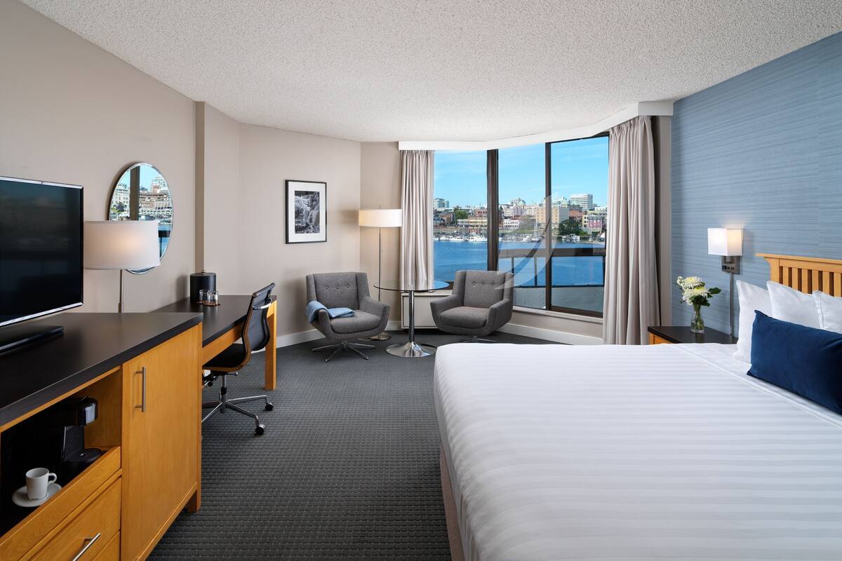 Habitación de hotel con cama, escritorio con televisor, dos sillones, una silla, lámpara, un cuadro en la pared y vista de la ciudad a través de amplias ventanas.