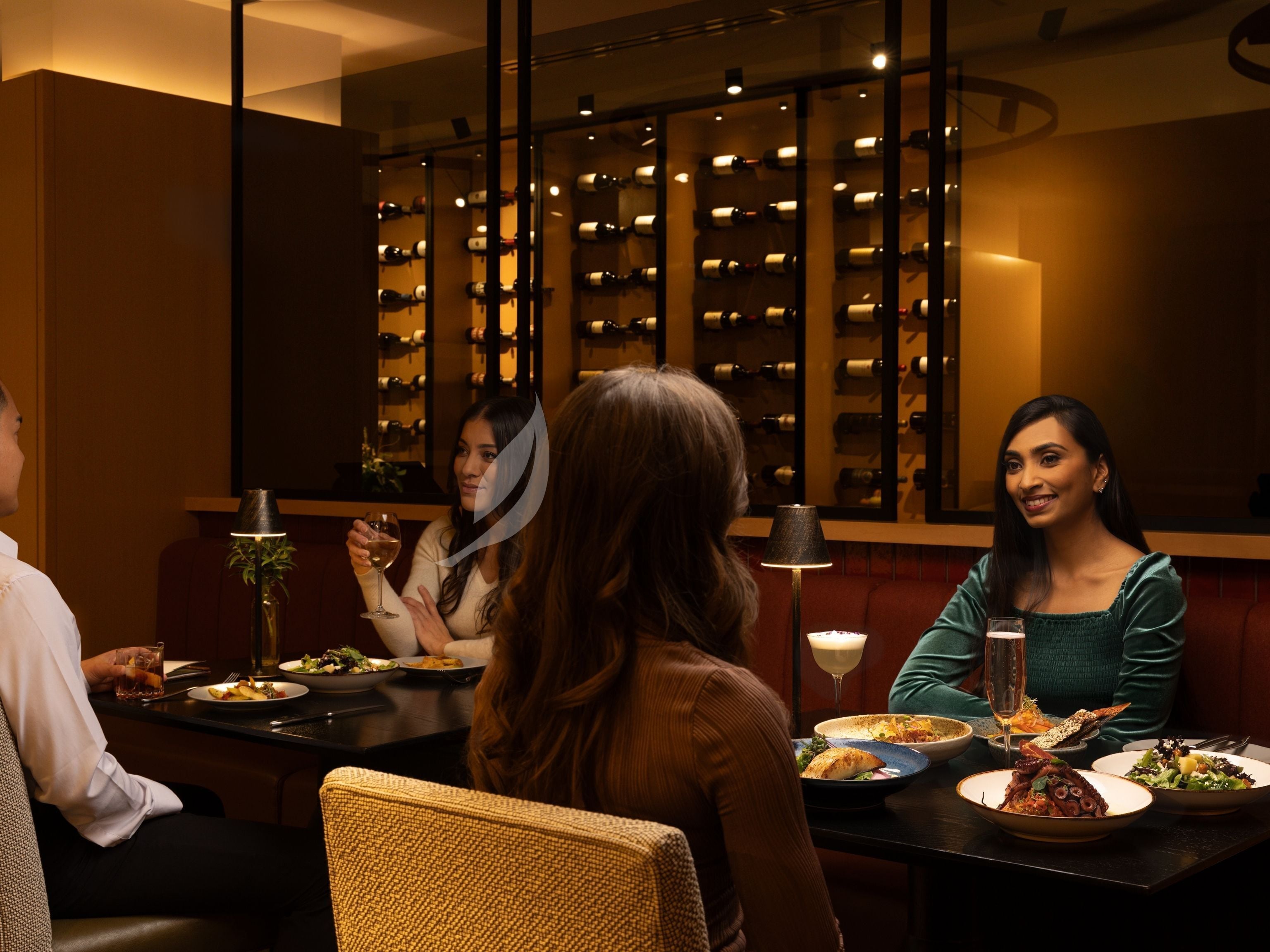 A restaurant with a wine rack in the background, a table with plates of food, and a lamp on the table.