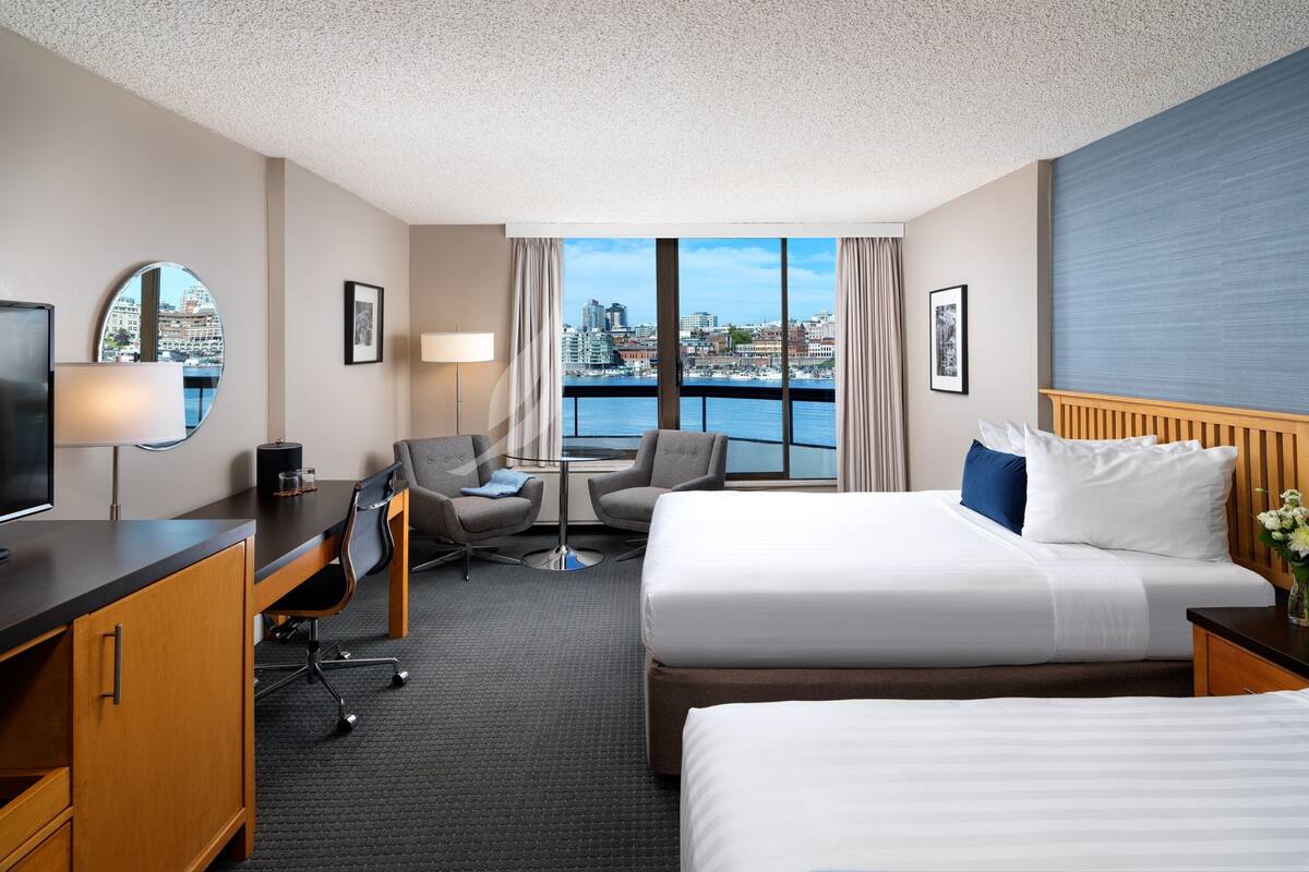 Habitación de hotel con cama, escritorio, silla, televisor y vista a la ciudad a través de amplias ventanas.
