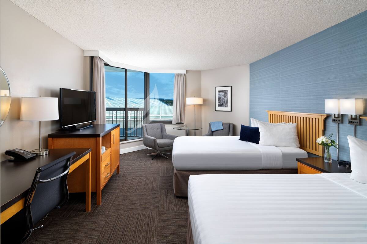 Habitación de hotel con cama, escritorio con televisor, silla, lámpara de escritorio y ventana con vista a un edificio.
