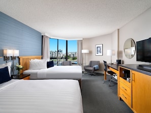 A hotel room with two beds, a desk, a chair, a TV, and a large window offering a city view.