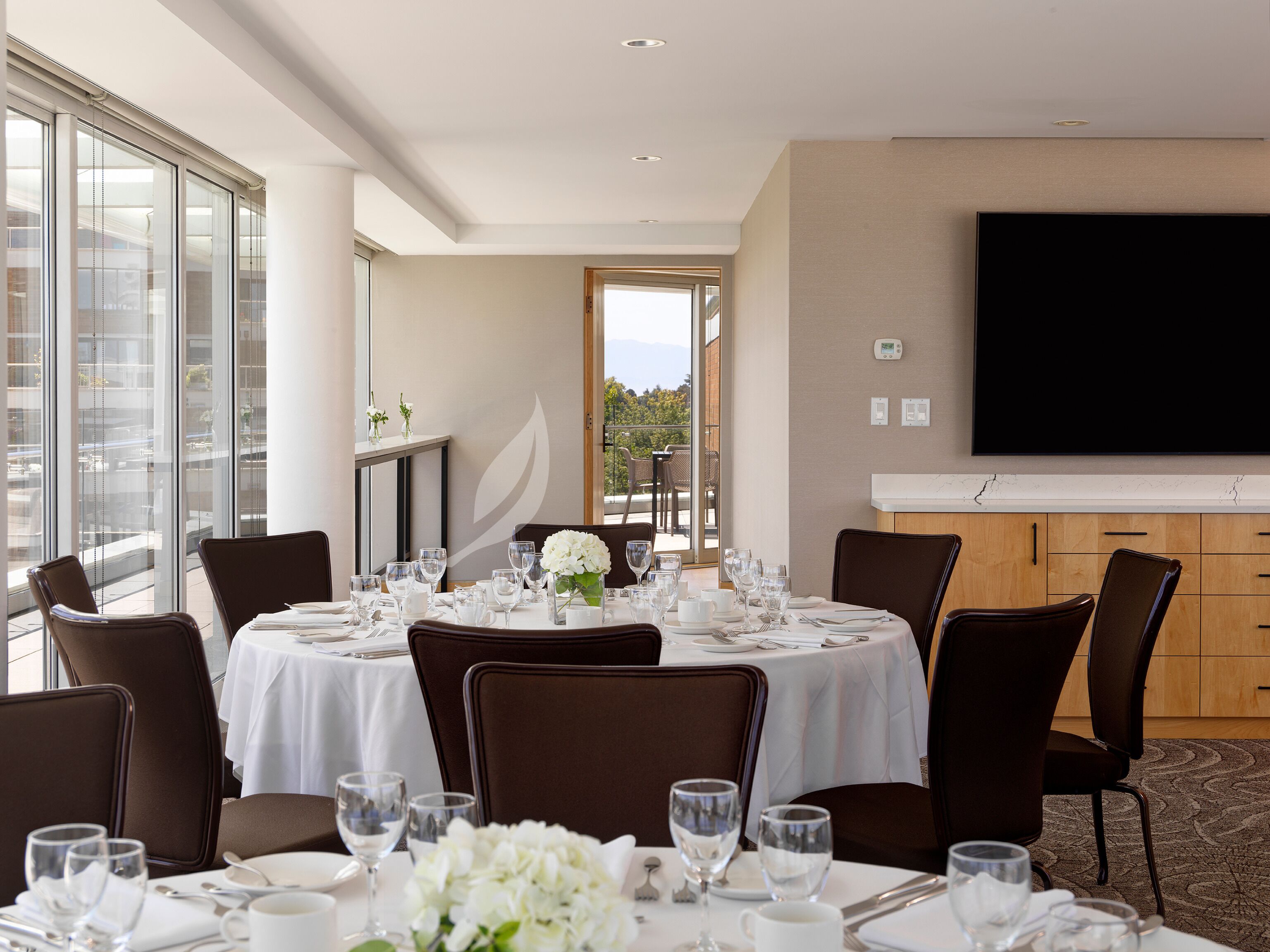 Un espace repas avec des tables rondes dressées pour le repas, de grandes fenêtres et un téléviseur à écran plat fixé au mur.