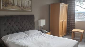 A hotel room with a tufted headboard bed, a wooden wardrobe, a bedside table with a lamp, and a view of the outdoors through a window.