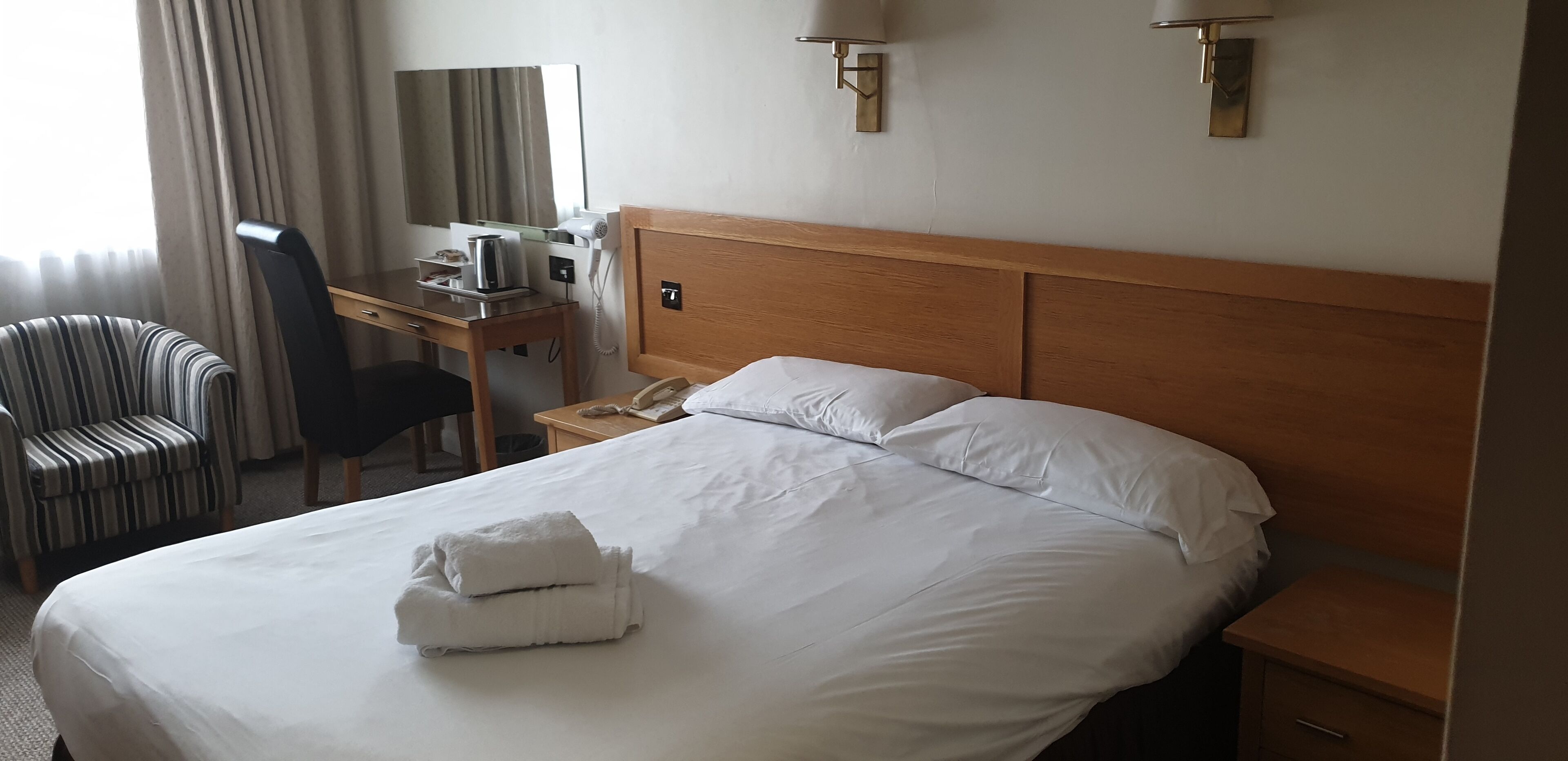A hotel room with a large bed, a desk with a telephone, a chair, and a lamp.