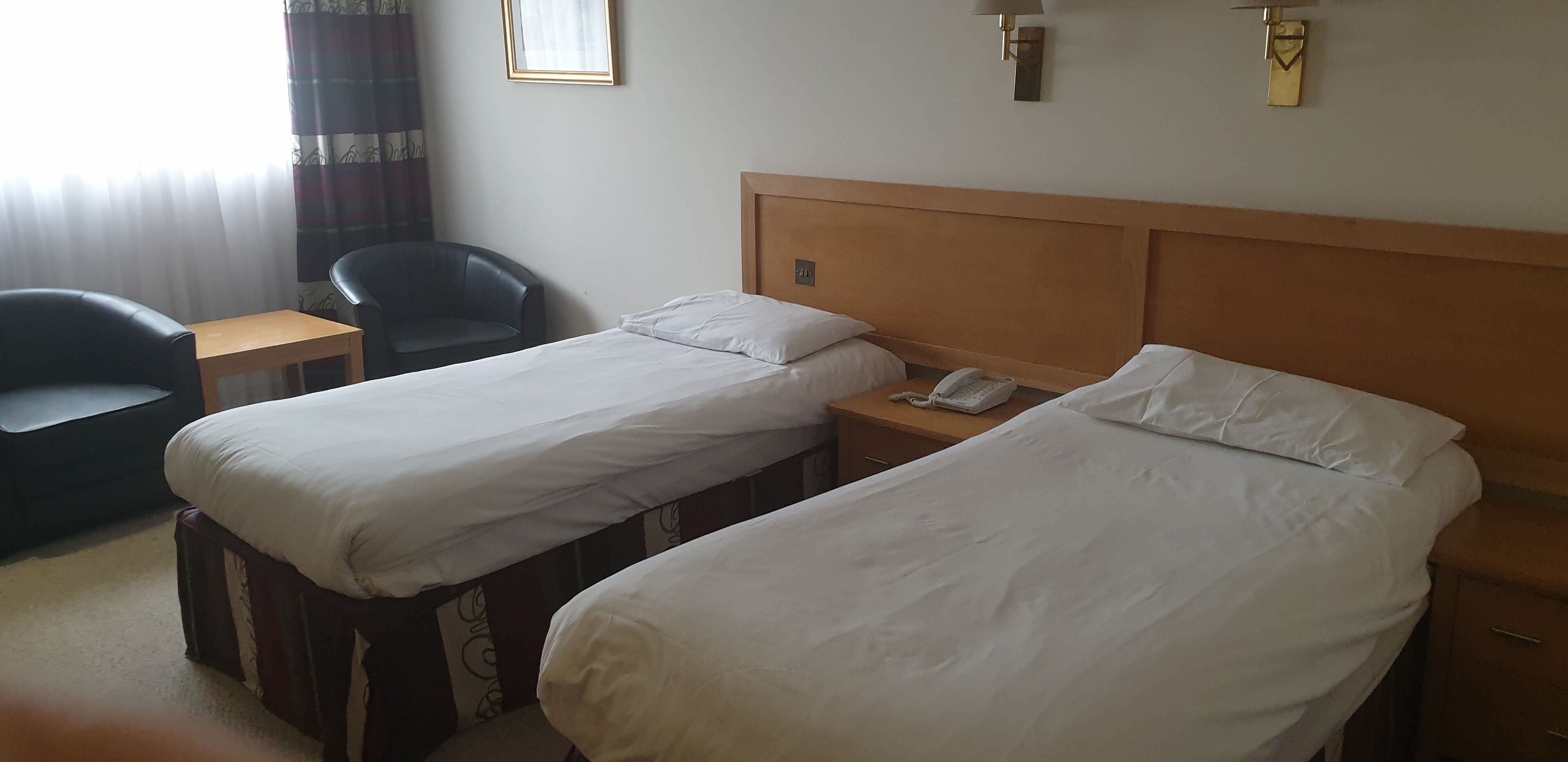 A hotel room with two single beds, a wooden headboard, a small wooden table, a black leather chair, and a telephone on the nightstand.