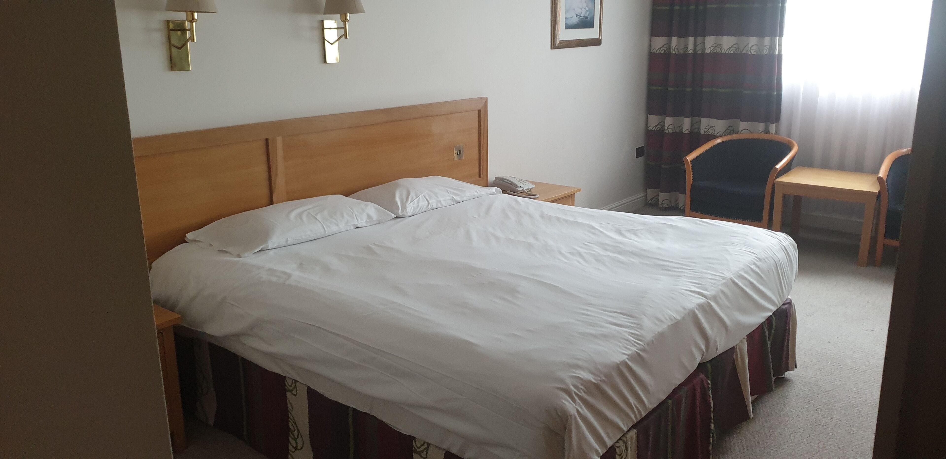 A hotel room with a large bed, a bedside table with a lamp, a chair, and a window with curtains.