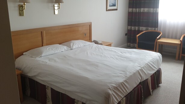 A hotel room with a large bed, a bedside table with a lamp, a chair, and a window with curtains.