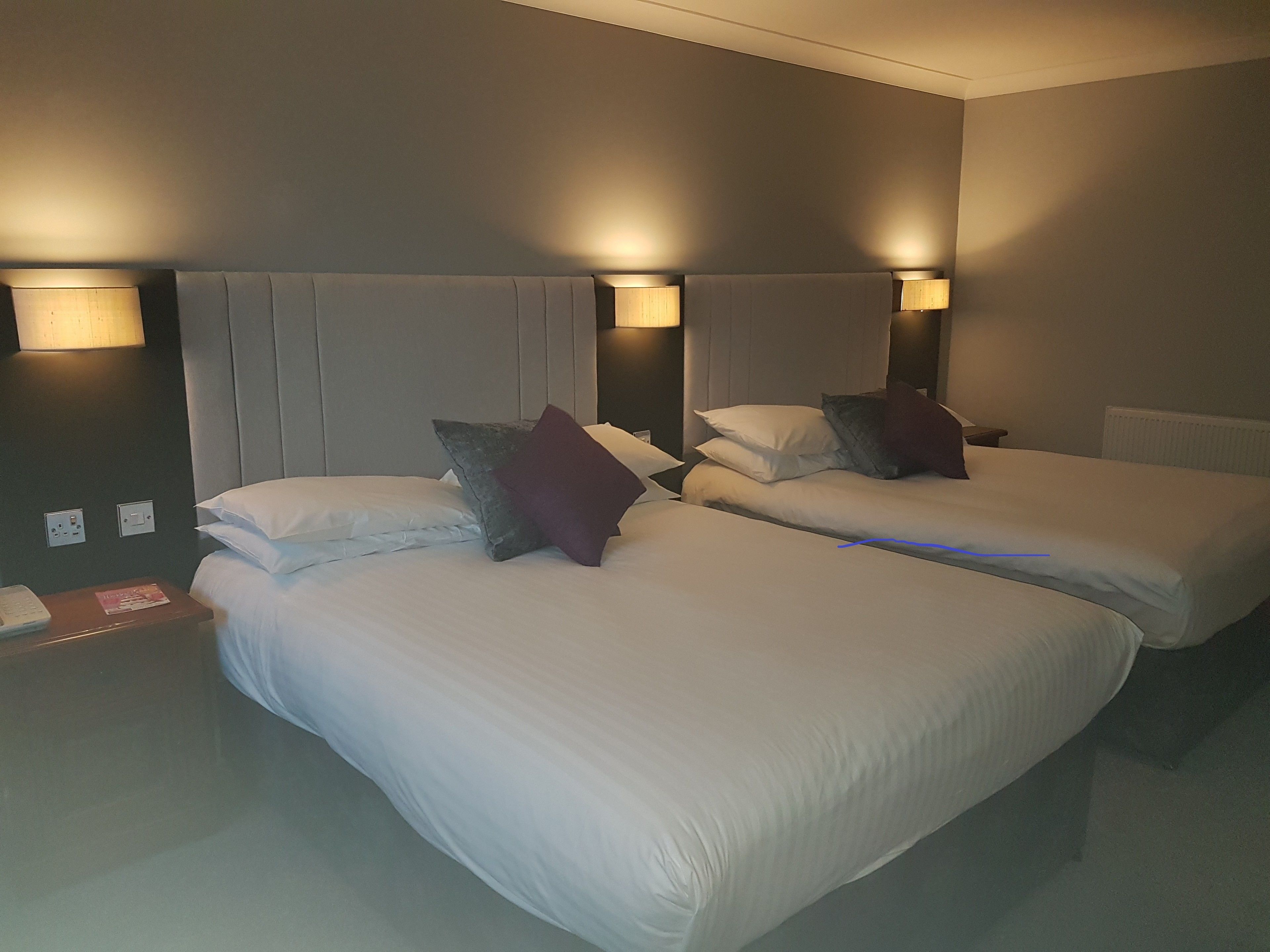 Habitación de hotel con dos camas individuales, cada una con sábanas blancas y una almohada. Hay lámparas de pared a cada lado de las camas.