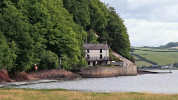 Une maison au bord d’une rivière, avec un quai, entourée d’une végétation luxuriante et avec des collines en arrière-plan.
